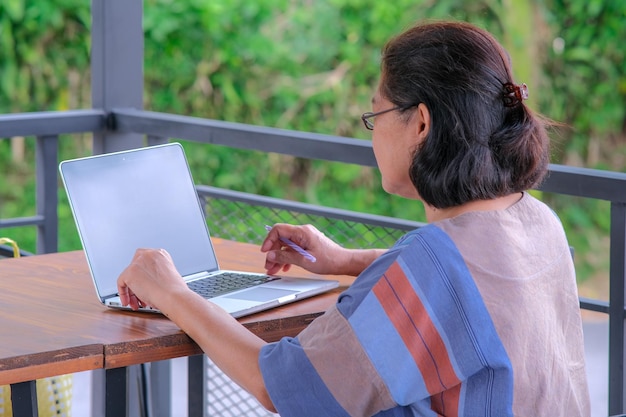 Mulher asiática sentada em sua varanda trabalhando em seu laptop com um copo de chá de limão na mesa