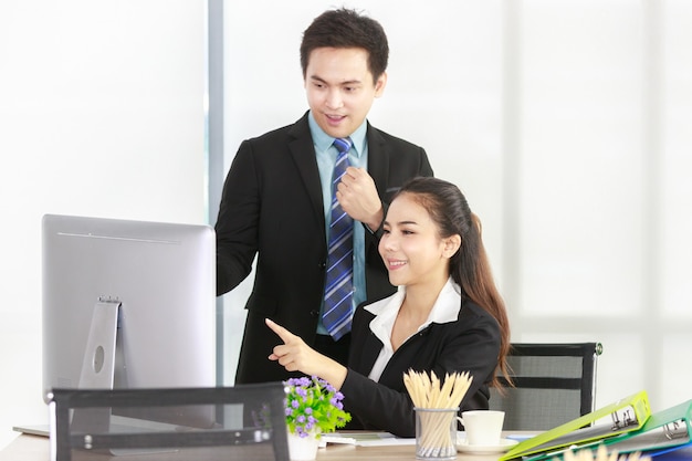 Mulher asiática sentada em uma mesa, apontando para um computador desktop e um empresário de pé feliz no trabalho no escritório. equipe de reunião de conceito para woirking.