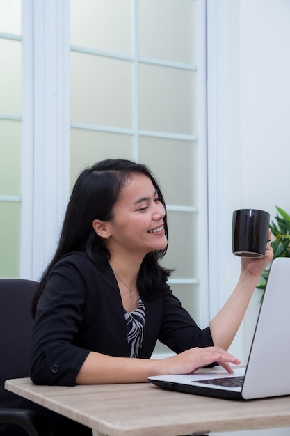 Mulher asiática sentada na cadeira enquanto sorrindo e segurando o copo na frente do laptop