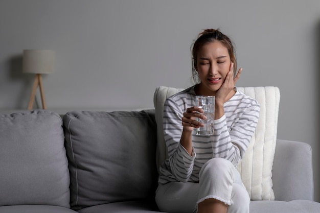 Foto mulher asiática sente dor de dente enquanto bebe água fria sentada no sofá em casa mulher com problemas de saúde dental