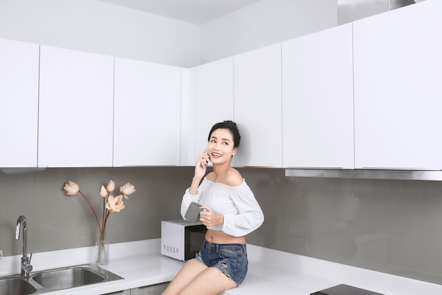 Mulher asiática sexy falando ao celular enquanto está sentado na cozinha.