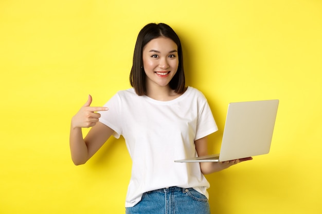 Mulher asiática sorridente apontando o dedo para seu laptop, mostrando algo online, em pé sobre um fundo amarelo