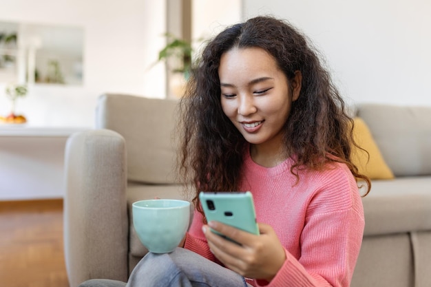 Mulher asiática sorridente atraente usando telefone inteligente enquanto está sentado no sofá em casa comunicação e conceito de chamada de vídeo