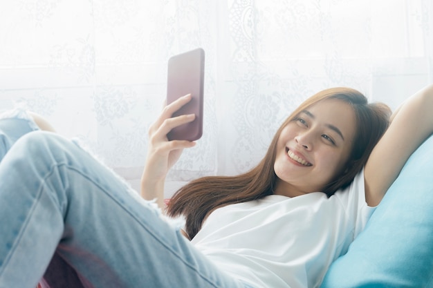Mulher asiática sorridente atraente usando telefone inteligente no sofá relaxante em casa.