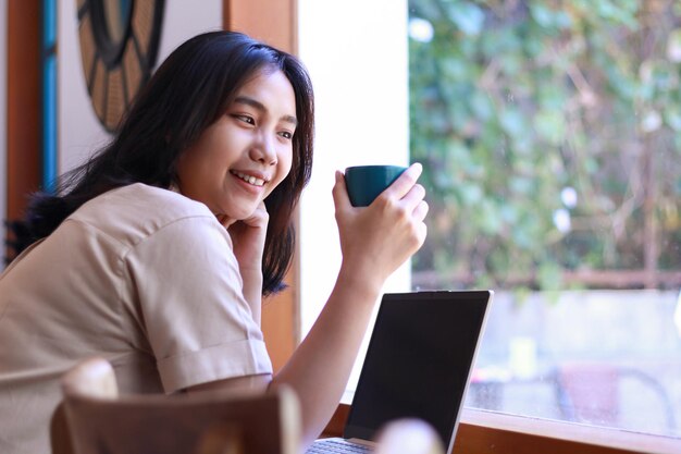 Mulher asiática sorridente bebendo café e usando laptop no espaço de trabalho do café vestindo roupas casuais e à moda
