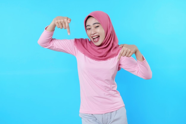 Mulher asiática sorridente com o dedo apontando isolado em um fundo azul claro usando hijab com camiseta rosa