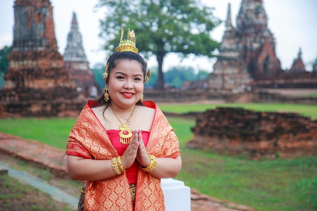 Mulher asiática sorridente em vestido tailandês com as mãos dobradas em saudação com fundo antigo templo de ruína com caminho de recorte