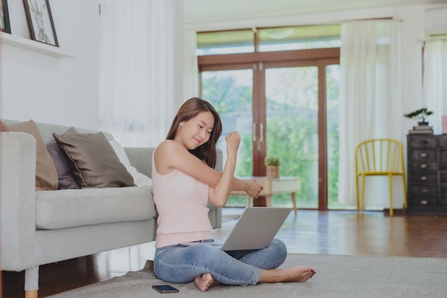 Mulher asiática sorridente esticando o ombro para relaxar depois de trabalhar em casa via laptop