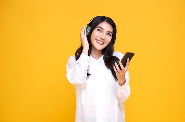 Mulher asiática sorridente feliz usando fones de ouvido sem fio, ouvindo música com o smartphone na parede amarela brilhante.