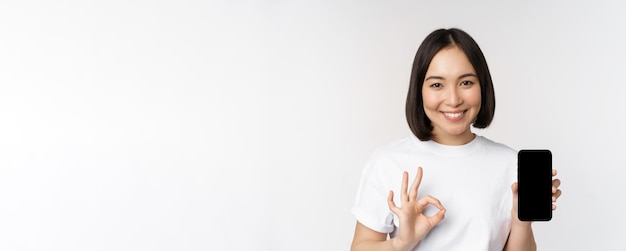 Mulher asiática sorridente, mostrando a tela do celular bem, recomendando o aplicativo de smartphone em pé sobre branco...