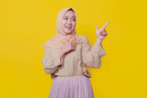 Mulher asiática sorridente usando hijab com o dedo apontando isolado no fundo do banner amarelo claro