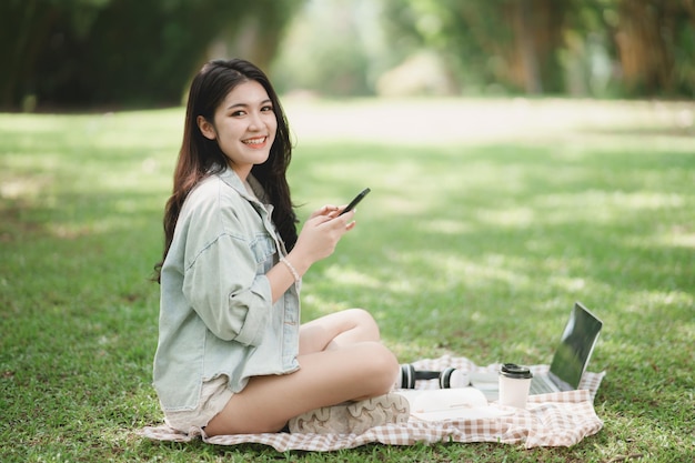 Mulher asiática sorrindo e sentado no cobertor de piquenique e gramado no parque usando telefone celular e trabalhando no laptop Mulher asiática usando compras móveis enquanto está sentado debaixo de uma árvore no parque Formulário de trabalho em qualquer lugar