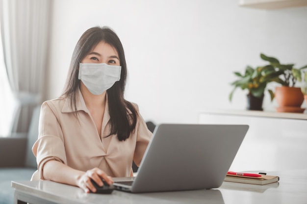 Mulher asiática sorrindo enquanto usava máscara facial trabalhando em casa com um laptop durante a quarentena de coronavírus