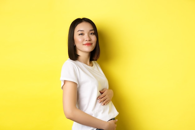 Mulher asiática, sorrindo gentilmente, tocando a barriga e olhando para a câmera, estar grávida, ter um bebê, em pé sobre fundo amarelo.