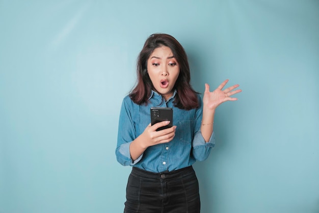 Mulher asiática surpreendida vestindo camisa azul apontando para seu smartphone isolado por fundo azul