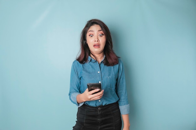 Mulher asiática surpreendida vestindo camisa azul apontando para seu smartphone isolado por fundo azul