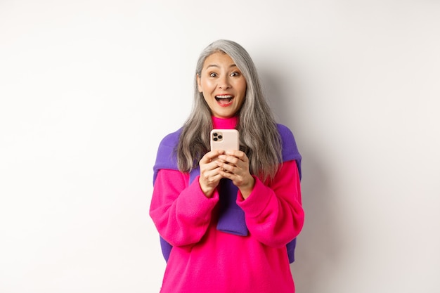 Mulher asiática surpresa, sorrindo depois de ler a promoção no smartphone, em pé com o celular em branco.