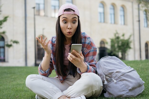 Mulher asiática surpresa usando um celular assistindo a um vídeo online sentada na grama