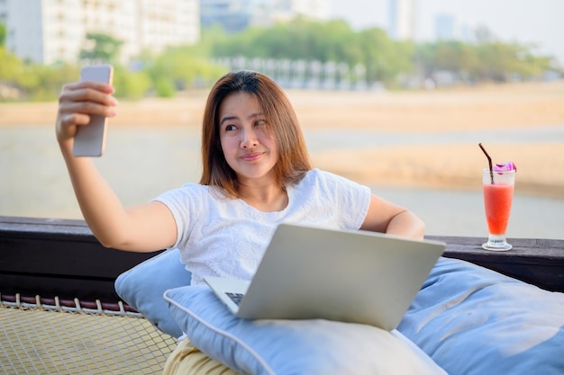 Mulher asiática tirando foto de selfie para mídias sociais no smartphone. Empresária trabalhando online na praia.