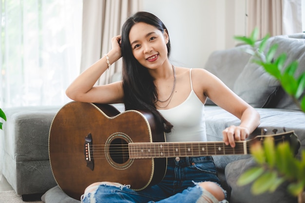 Mulher asiática tocando violão em casa, estilo de vida de jovem guitarrista músico com instrumento de arte acústica sentada para tocar e cantar uma música fazendo som em hobby na sala de casa