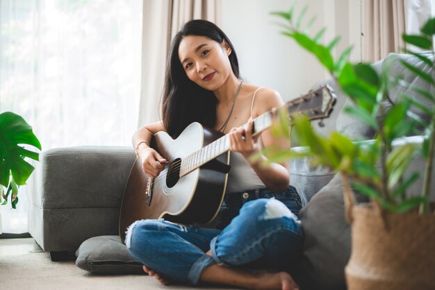 Mulher asiática tocando violão em casa, estilo de vida de jovem guitarrista músico com instrumento de arte acústica sentada para tocar e cantar uma música fazendo som em hobby na sala de casa