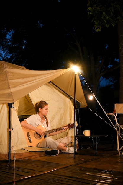 Mulher asiática tocando violão na frente da barraca no acampamento à noite