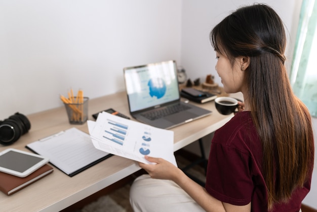 Mulher asiática trabalhando com laptop e documento enquanto está sentada no escritório da sala de estar como freelancer