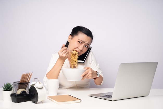 Mulher asiática trabalhando duro horas extras comendo macarrão instantâneo no escritório. Empresária chateada, excesso de trabalho e fome ocupada.