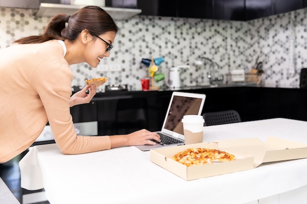 Mulher asiática trabalhando em casa na cozinha e comendo pizza no delivery