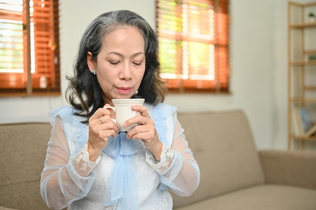 Mulher asiática tranquila e relaxada de 60 anos tomando chá da tarde em sua sala de estar