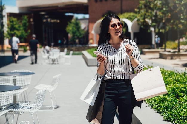 mulher asiática usa óculos de sol em pechincha verão venda alegremente comprando na loja de roupas. Moça feliz com sacolas de compras, gostando de passear no shopping em stanford. Conceito de estilo de vida de compra de consumismo