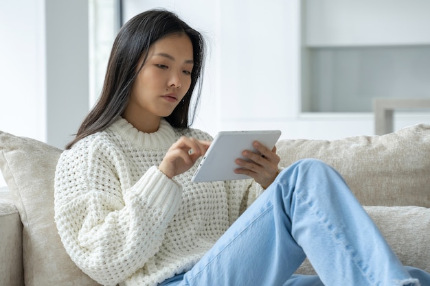 Mulher asiática usa um tablet enquanto está sentada em um sofá em sua sala de estar, uma mulher feliz usa um