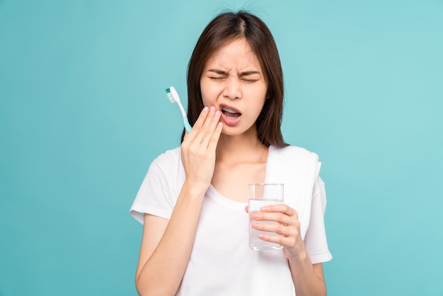 Mulher asiática usando aparelho com escovar os dentes com dentes sensíveis e segurando o copo de água, toalha no ombro sobre fundo azul, conceito de higiene oral e cuidados de saúde.