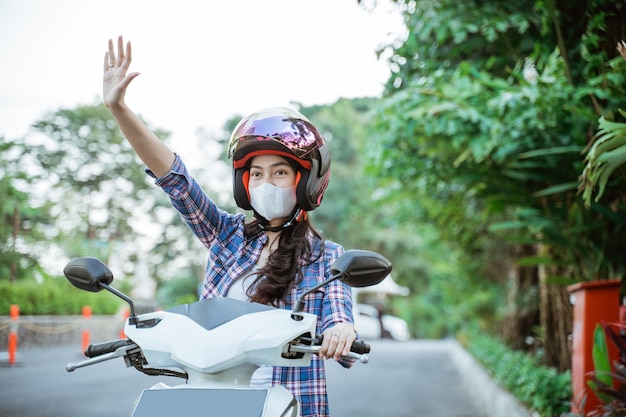 Mulher asiática usando capacete e máscara acenando enquanto andava de moto na rua