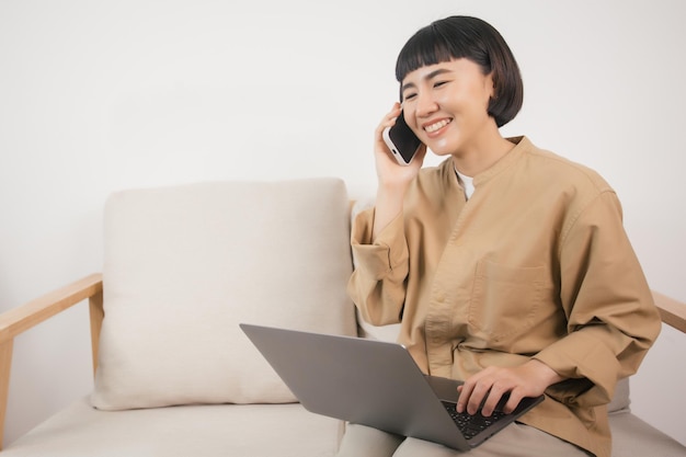 Foto mulher asiática usando laptop enquanto está sentado no sofá em casa e falando ao telefone