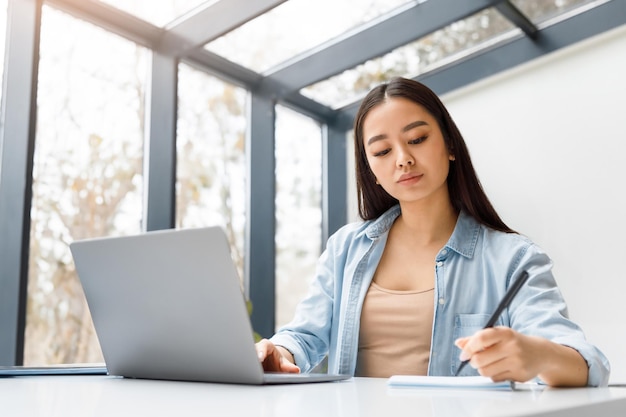 Mulher asiática usando laptop surfando na web e tomando notas sentada em uma mesa em um escritório de coworking ou
