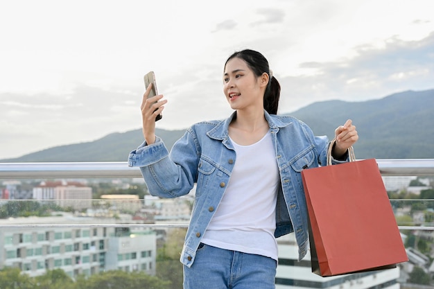 Mulher asiática usando seu smartphone carregando suas sacolas de compras em pé no telhado do shopping da cidade