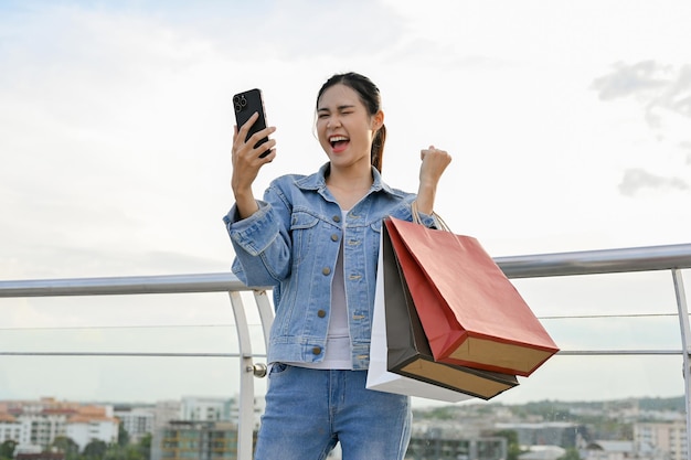 Mulher asiática usando smartphone enquanto carrega sacolas de compras gosta de fazer compras