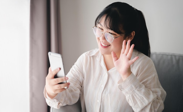Mulher asiática usando smartphone para videoconferência com amigos acenando com a mão fazendo gesto de olá
