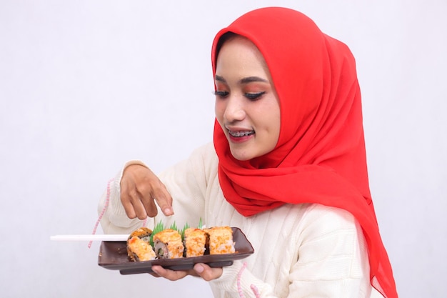 Mulher asiática usando um hijab em ângulo reto alegre e sincera gestando para mostrar sushi segurando