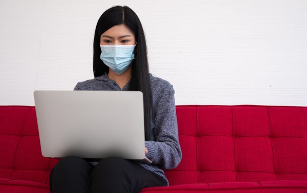 Mulher asiática usando uma máscara facial e usando um laptop no sofá para trabalhar em casa