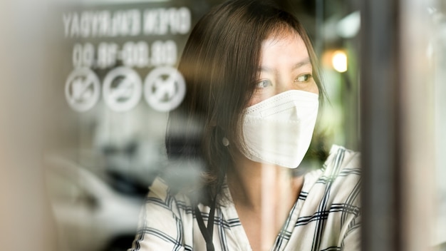 Mulher asiática usando uma máscara médica, olhando pela janela de um café, com os olhos tristes, trabalhando em casa, quarentena.