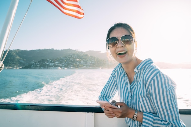 mulher asiática viajando na moda está rindo da câmera enquanto estiver usando smartphone pelo trilho em um ferry boat durante as férias de verão na Califórnia EUA com luz de fundo
