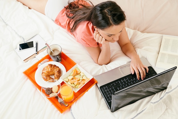 Mulher assistindo o computador portátil e tomando café da manhã. Ela está no quarto. Visão aérea