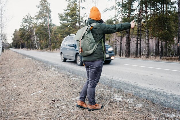 Mulher ativa com mochila caminhando na estrada e gesto tentando parar o carro, pedindo carona no turismo solo