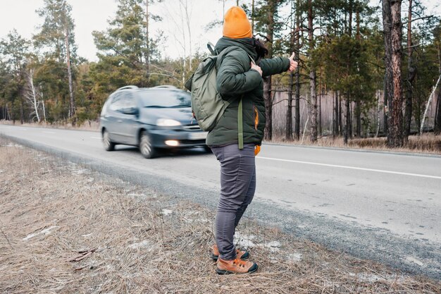 Mulher ativa de meia idade com mochila caminhando na estrada e gesto tentando parar um carro, fundo de floresta, conceito de viagens de inverno, estilo de vida saudável, turismo solo de carona