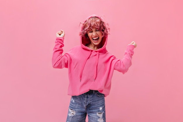 Mulher ativa feliz com capuz rosa e calças jeans rindo e dançando em fundo isolado Retrato de jovem legal de bom humor no pano de fundo rosa