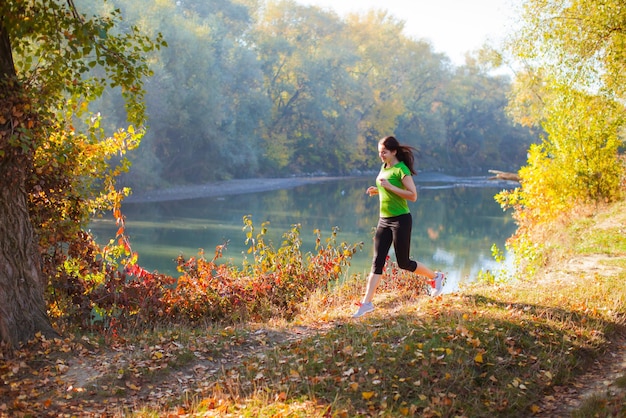 Mulher ativa slimfit treinando no parque da cidade outono perto do rio. Mulher com conceito de estilo de vida saudável fazendo sua rotina de corrida no parque da cidade outono. Floresta de outono ensolarada ao fundo
