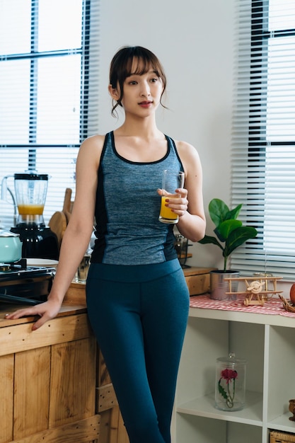 mulher atlética asiática de tiro vertical está de pé com elegância e olhando para a distância enquanto toma suco fresco em uma manhã tranquila após o treino em casa.