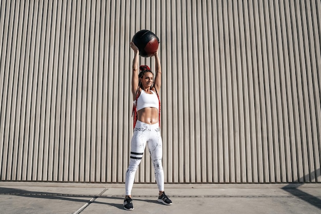 Mulher atlética de altura total fazendo exercício com bola médica aumenta sobre sua cabeça Força e motivaçãoFoto de mulher esportiva em roupas esportivas da moda
